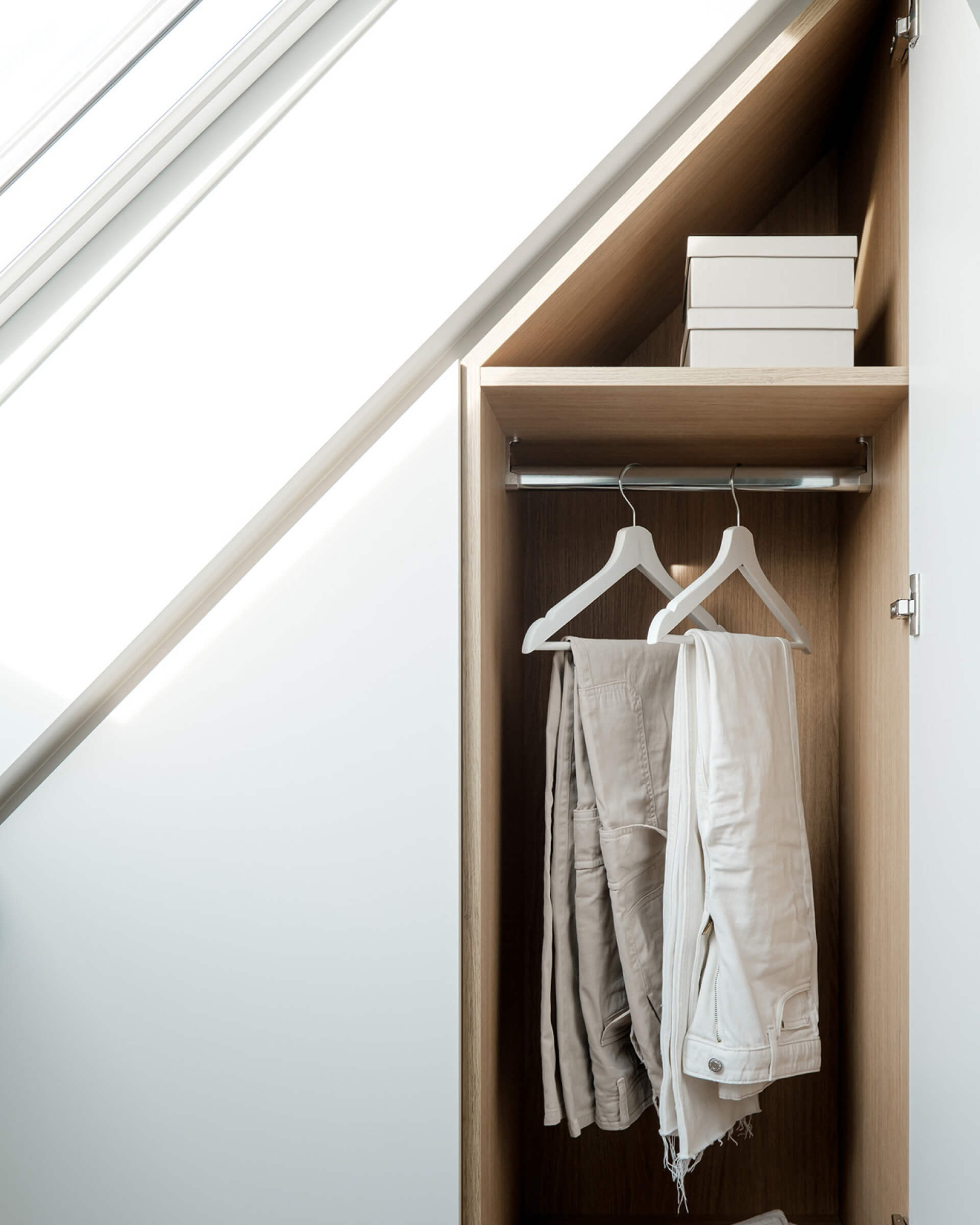 Des cintres blancs portent deux pantalons dans une armoire blanche sous un toit en pente.