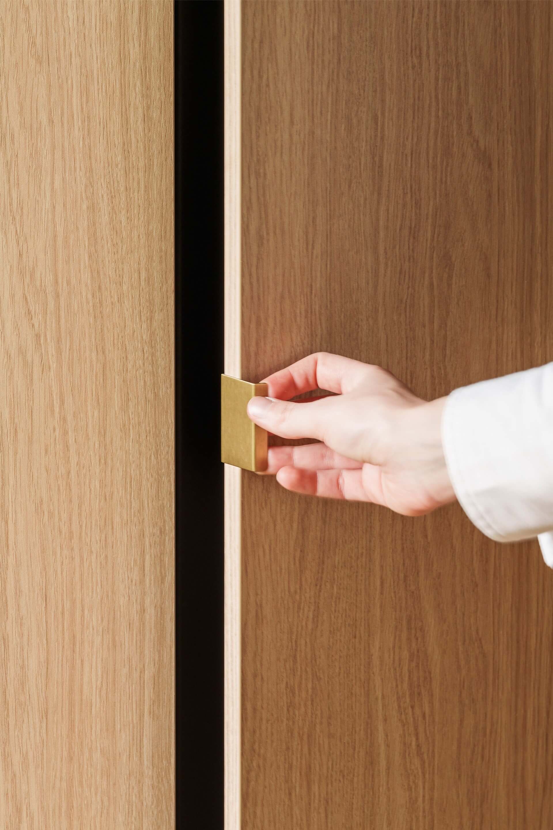 Gros plan sur une poignée Mini Pure en laiton massif fixée sur un meuble sur mesure en Atlas Oak. À droite, une main ouvre la porte du meuble.