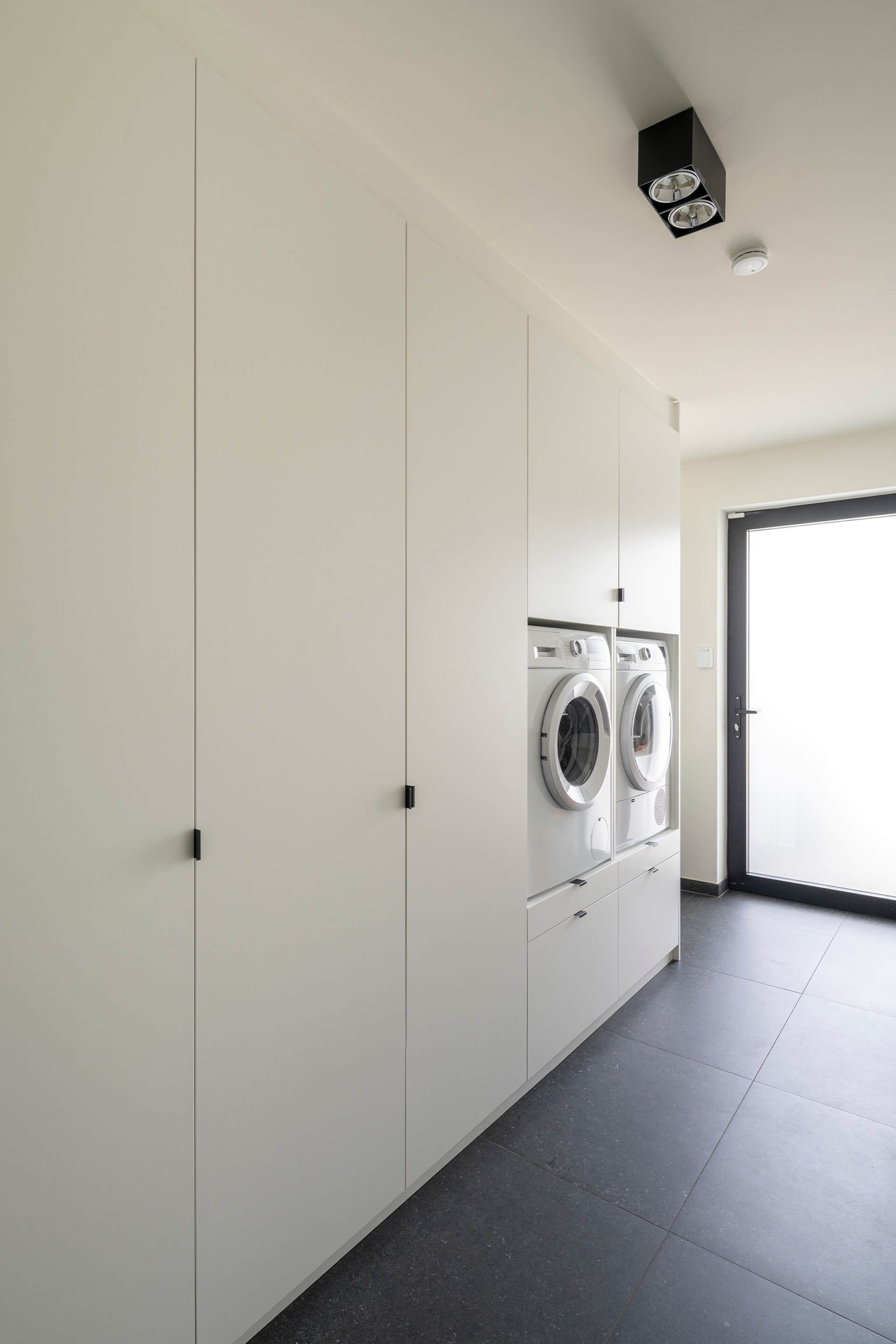 Meuble de buanderie blanc avec poignées Mini Pure noires. À l'extrême droite de la photo, on aperçoit une femme rangeant du linge dans un placard au-dessus de la machine à laver.