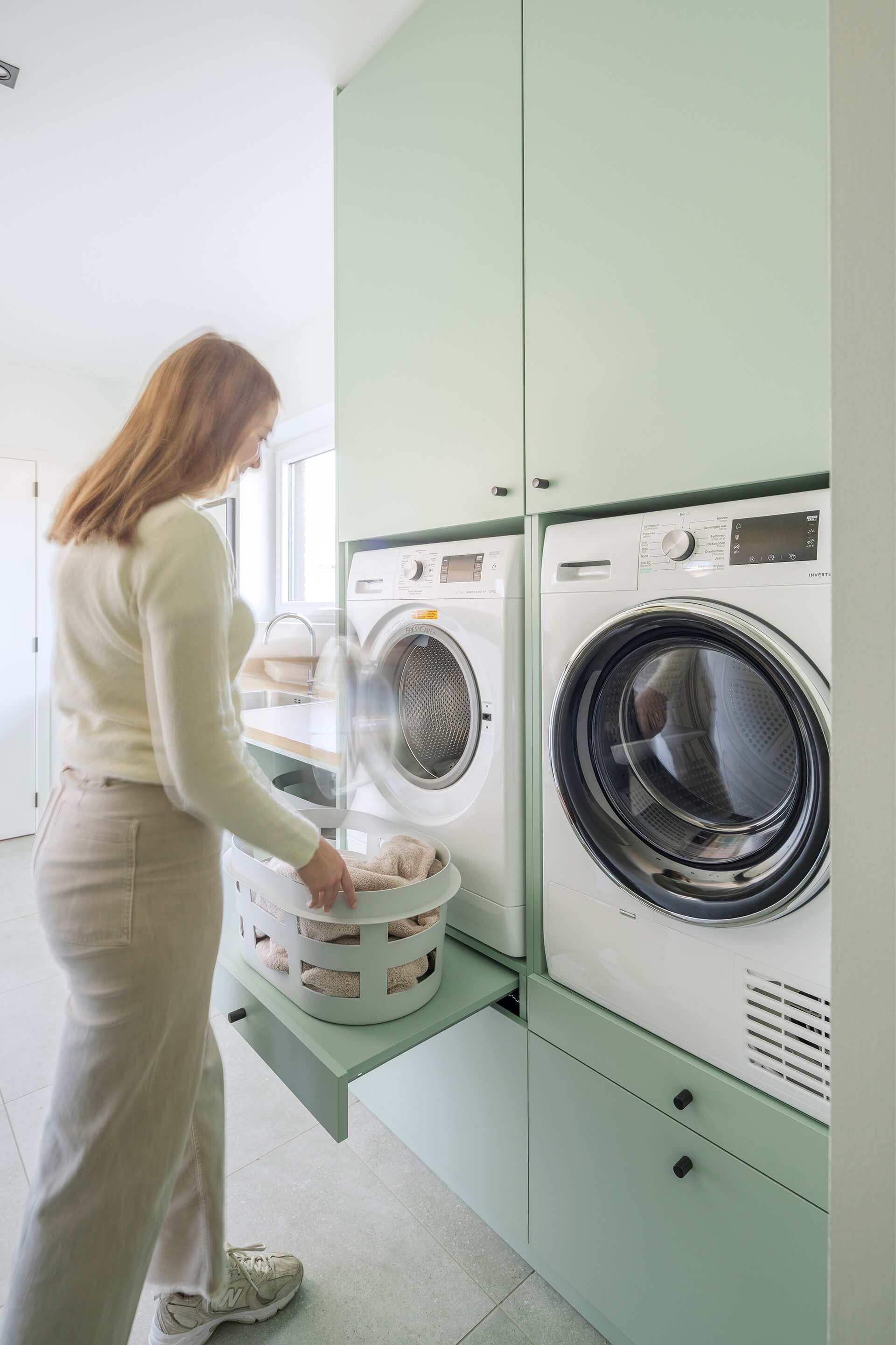 Meuble de buanderie sur mesure, de couleur Industrial Green avec poignées Monument noires. À gauche de la photo, on aperçoit une femme ouvrant une machine à laver.