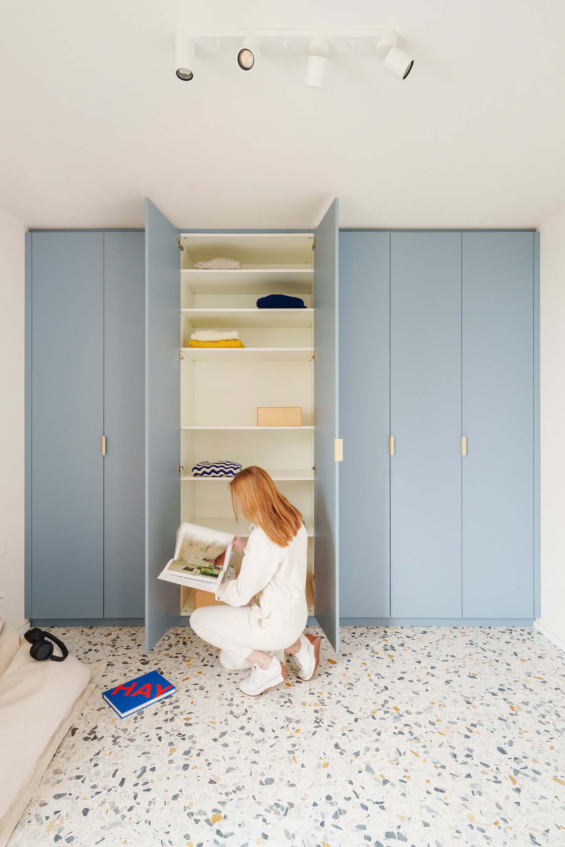 Armoire sur mesure en Silver Blue avec poignées tonton crème. La porte centrale est ouverte. En diagonale devant l'armoire, une femme est assise sur un pouf bas et lit des magazines.