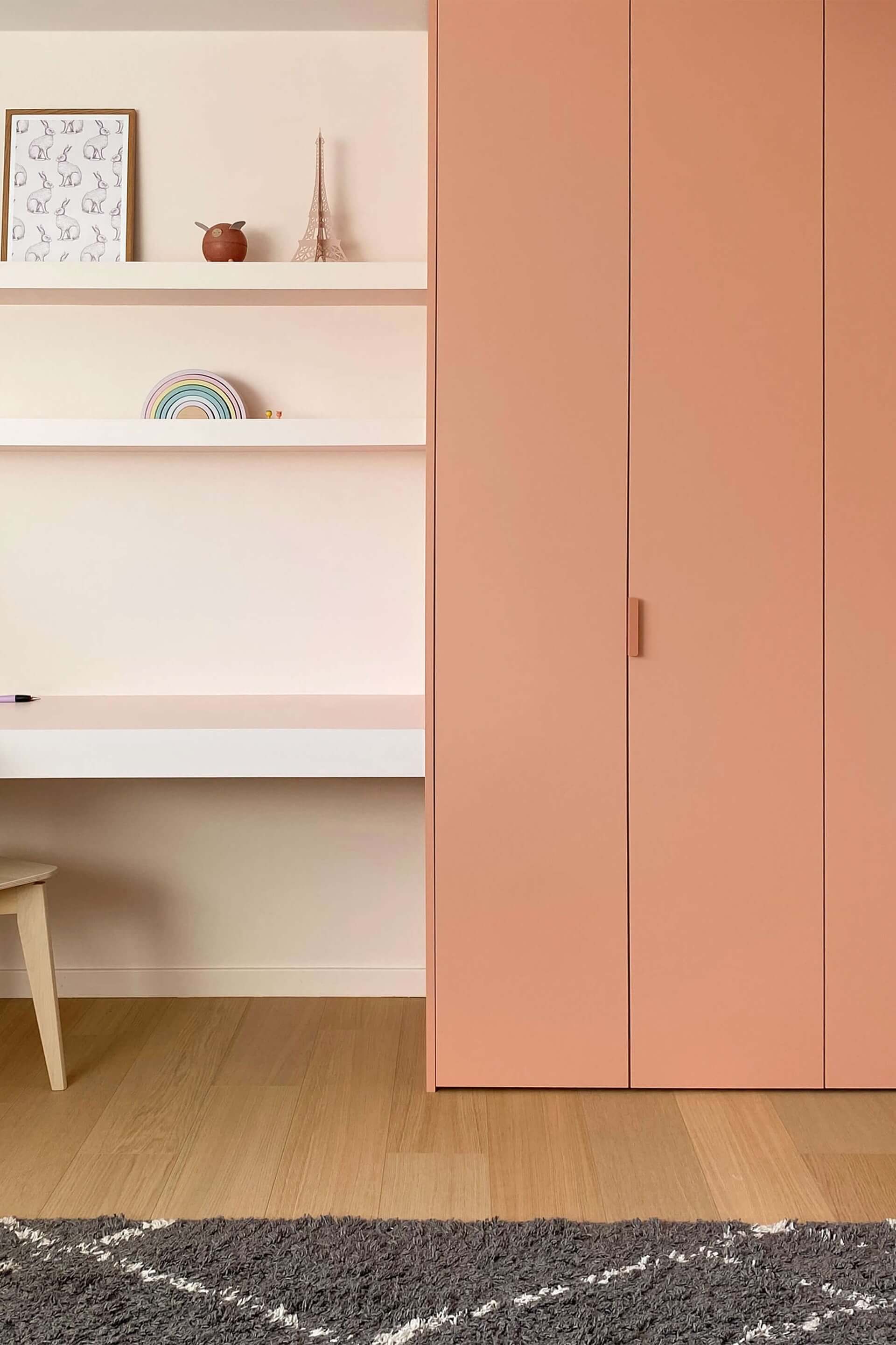 Armoire de chambre d'enfant sur mesure, couleur corail poudré, avec poignées Tonton roses. L'armoire se trouve à droite sur la photo, un bureau et une chaise en bois étant à gauche.